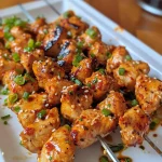 Close-up of juicy Bang Bang Chicken Skewers on a wooden platter, garnished with green onions and sesame seeds.
