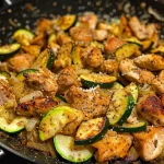 Close-up of a sizzling chicken and zucchini skillet with golden-brown pieces of chicken and vibrant green zucchini.
