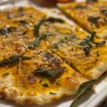 ['Close-up of a golden brown pumpkin and fried sage flatbread with crispy edges and melted cheese.', 'Side view of a freshly baked flatbread topped with pumpkin, fried sage leaves, and Parmesan cheese.', 'Appetizing flatbread featuring pumpkin and sage, garnished with a drizzle of honey and fresh herbs.', 'Deliciously arranged flatbread showcasing roasted pumpkin and crunchy sage leaves on a wooden surface.']